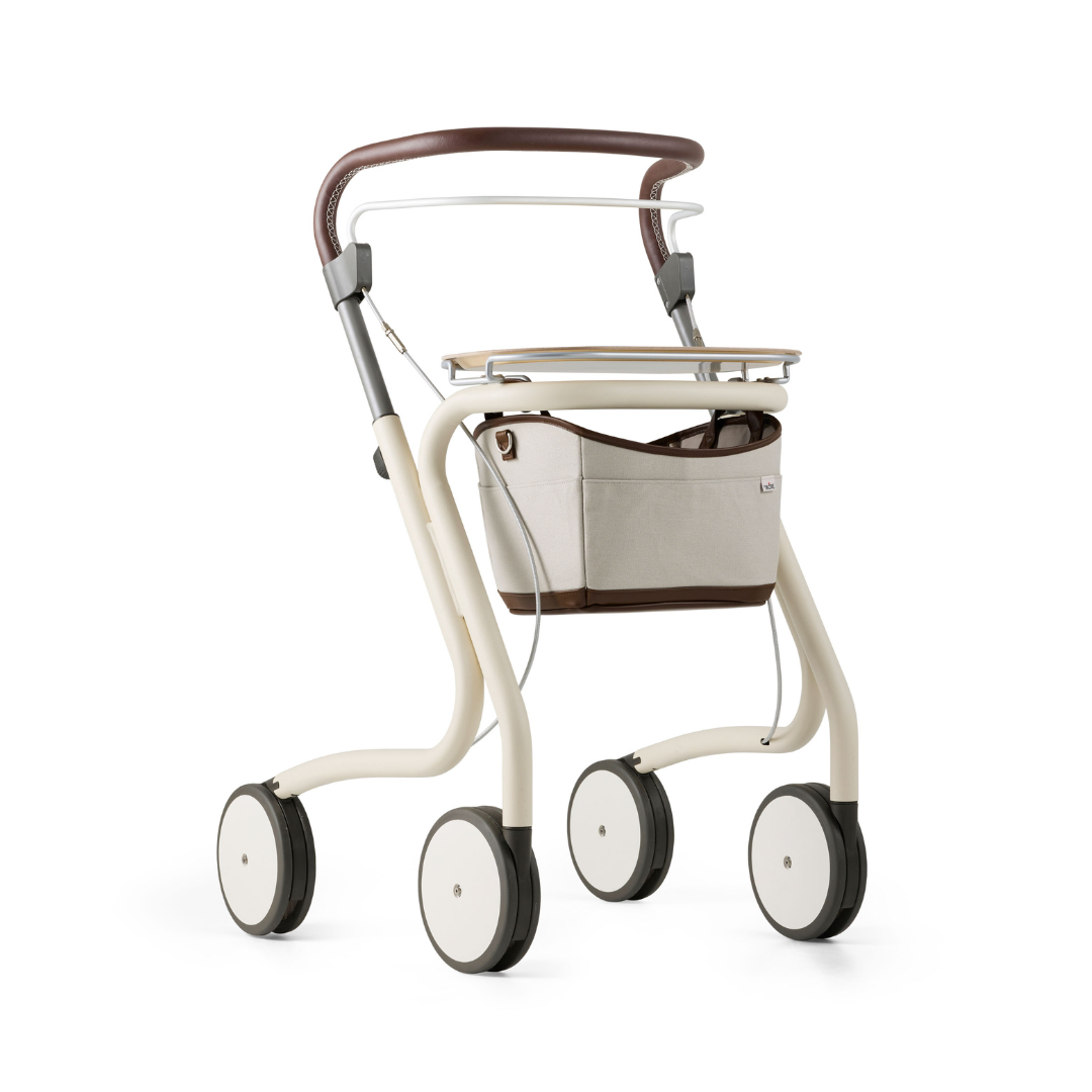 The byACRE Scandinavian Butler At-Home Rollator features a cream frame, brown handles, and a beige storage bag below the seat. This modern, four-wheeled rollator offers sleek, minimalist Scandinavian design for stylish at-home mobility.